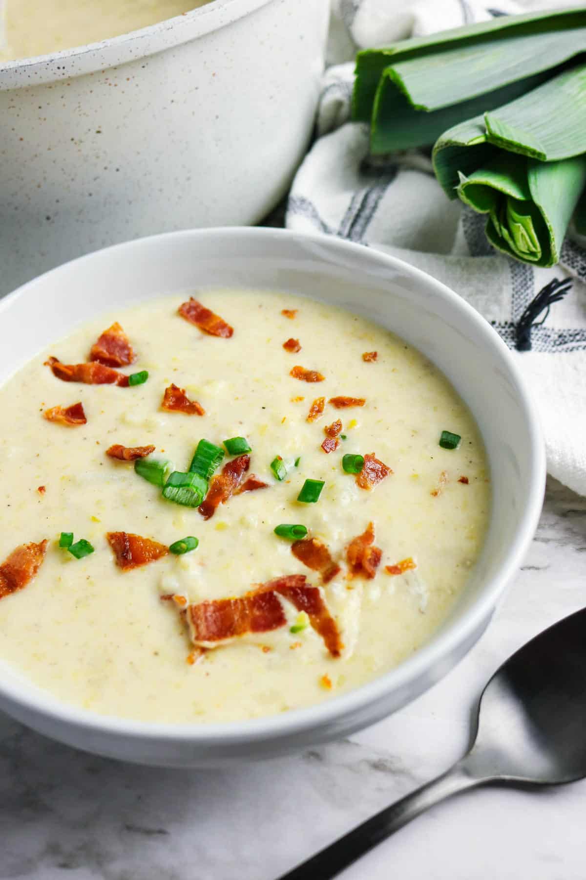 A white bowl of creamy bacon potato and leek soup, topped with chopped bacon and green onions, sits on a marble surface near a spoon, a checkered towel, and fresh leeks.