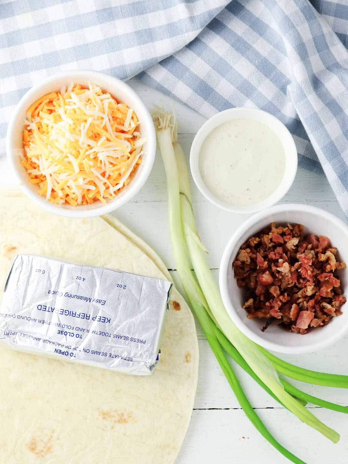 A tortilla, cream cheese, shredded cheese, bacon bits, green onions, and a cup of ranch dressing—perfect ingredients for delicious bacon ranch pinwheels—sit on a white wooden surface with a blue and white checkered cloth.