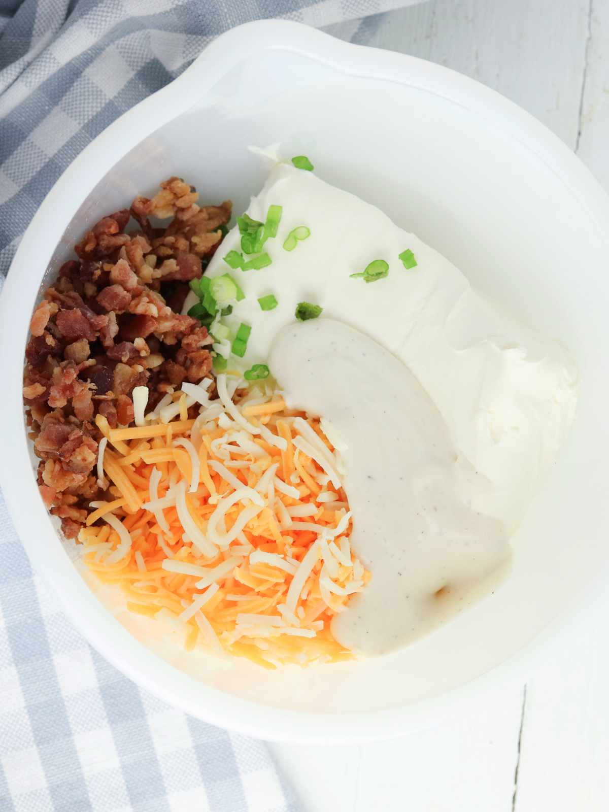 A white bowl containing chopped bacon, shredded cheese, sliced green onions, cream cheese, and a dollop of ranch dressing-perfect for making delicious bacon ranch pinwheels-sits on a gray and white checkered cloth.