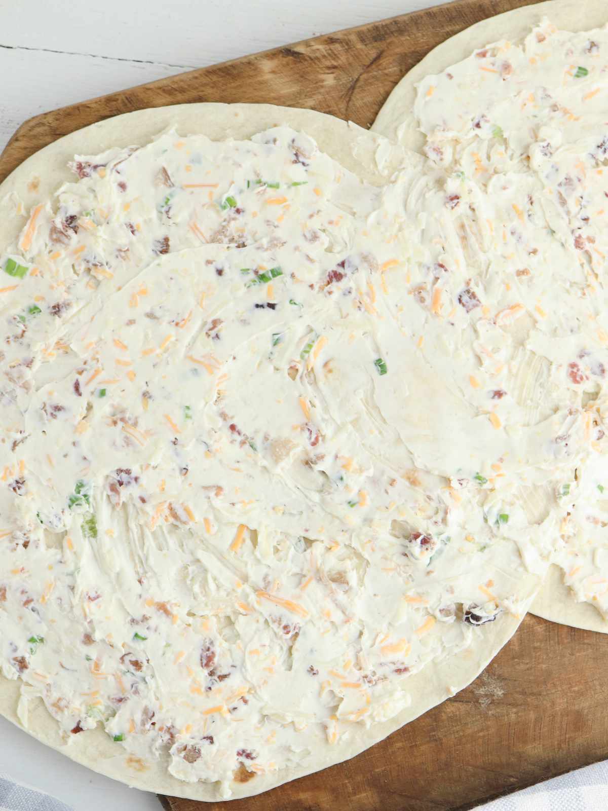 Two large flour tortillas on a wooden board, spread with a creamy mixture of bacon, shredded cheese, and chopped green onions-perfect for making delicious bacon ranch pinwheels.