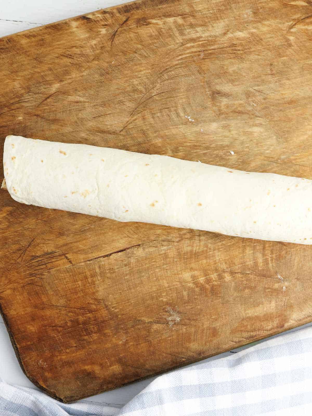 A rolled flour tortilla, ready to become delicious bacon ranch pinwheels, sits on a rustic wooden cutting board, with a checkered white and gray cloth visible in the bottom corner.