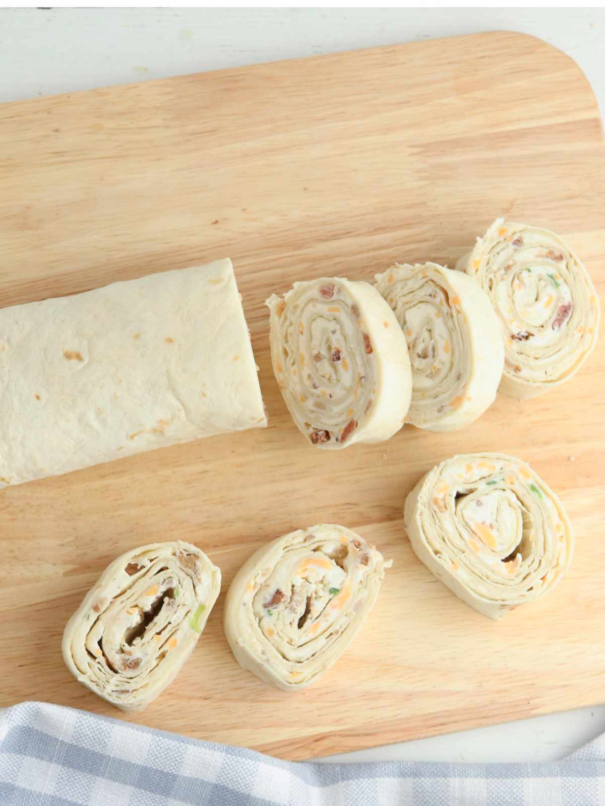 A partially sliced tortilla wrap on a wooden cutting board, showing several bacon ranch pinwheels with a creamy filling and bits of green and brown. A striped cloth is partially visible in the bottom corner.