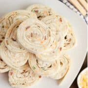 A plate of neatly stacked bacon ranch pinwheels filled with a creamy mixture containing shredded cheese and green herbs, arranged in a spiral pattern on a white dish.