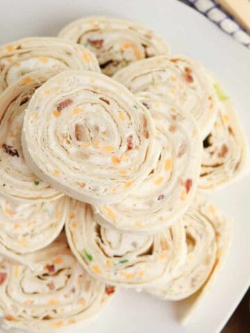 A plate of neatly stacked bacon ranch pinwheels filled with a creamy mixture containing shredded cheese and green herbs, arranged in a spiral pattern on a white dish.