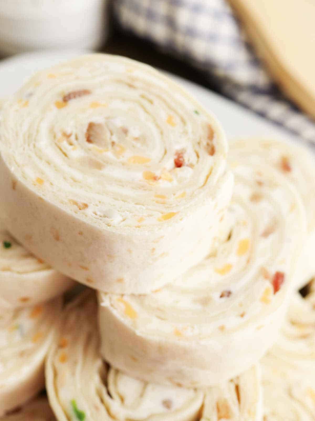 Close-up of several crack pinwheels filled with a creamy mixture, bits of cheese, and green herbs, stacked on a plate. A checkered cloth is blurred in the background.