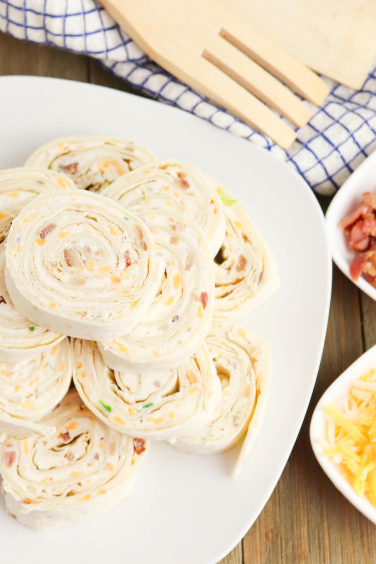 A white plate with several bacon ranch pinwheels, filled with cream cheese, herbs, and bacon bits. The plate is on a wooden table next to shredded cheese, bacon, and a wooden spatula.
