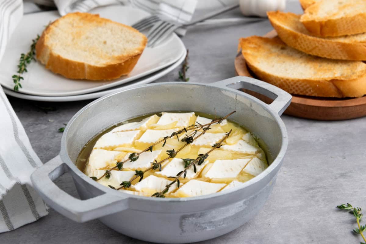 A round baking dish filled with baked brie with honey and herbs, next to toasted bread slices on a wooden plate and a stack of white plates with utensils on a gray surface.