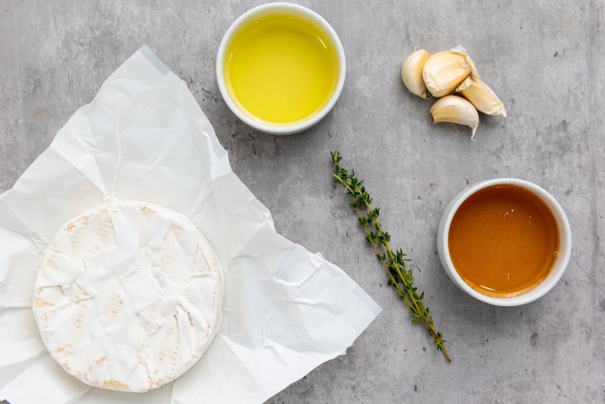 A round of brie cheese on white parchment, ready for baked brie with honey, sits beside a small bowl of olive oil, a bowl of honey, three garlic cloves, and a sprig of fresh thyme on a gray surface.