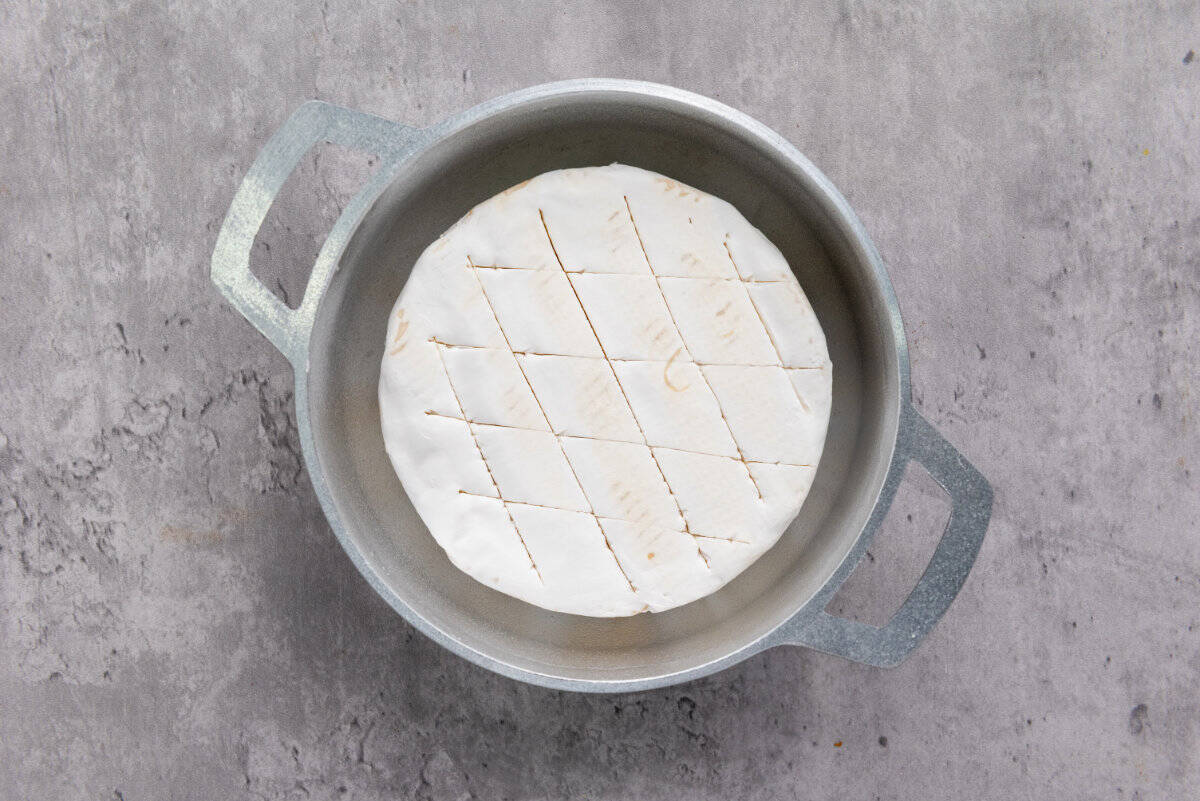 A round block of white cheese with a grid pattern cut into the surface sits in a metal pot on a gray countertop, ready to be transformed into baked brie with honey.