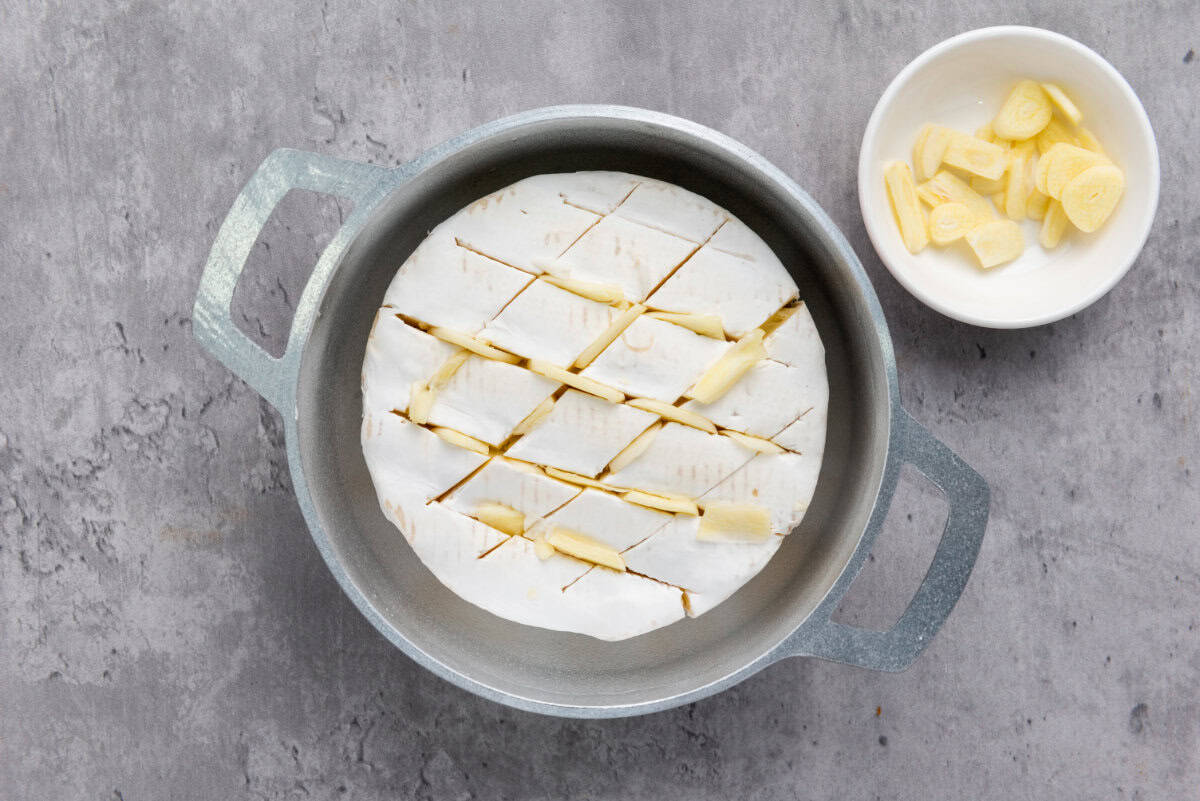 A round wheel of baked brie with honey sits in a metal baking dish, its top scored with crisscross cuts and filled with slices of garlic. A small white bowl holding extra garlic slices rests nearby on the gray surface.