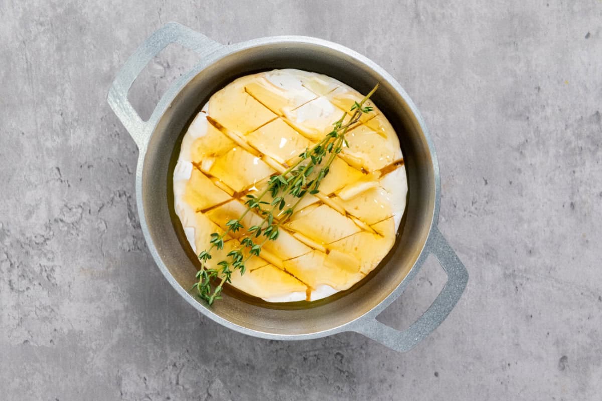 A wheel of baked brie with honey, scored in a crisscross pattern and topped with fresh thyme sprigs, sits in a small metal baking dish on a gray surface.