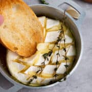 A round of baked brie with honey, topped with herbs and garlic in a small metal dish, with a slice of toasted bread being dipped into the gooey melted cheese.