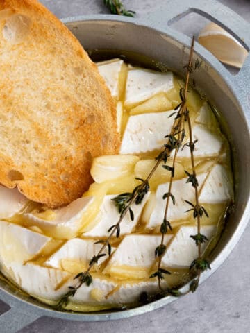 A round of baked brie with honey, topped with herbs and garlic in a small metal dish, with a slice of toasted bread being dipped into the gooey melted cheese.