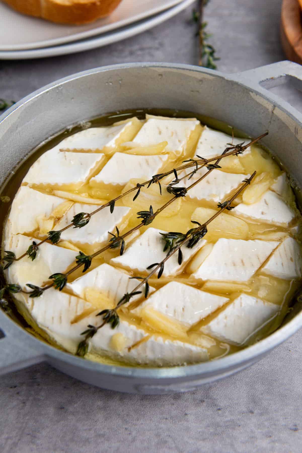 A round wheel of brie cheese scored with a diamond pattern, topped with fresh thyme and drizzled with honey, then baked in a small gray dish until perfectly melted—this baked brie with honey is an irresistible appetizer.