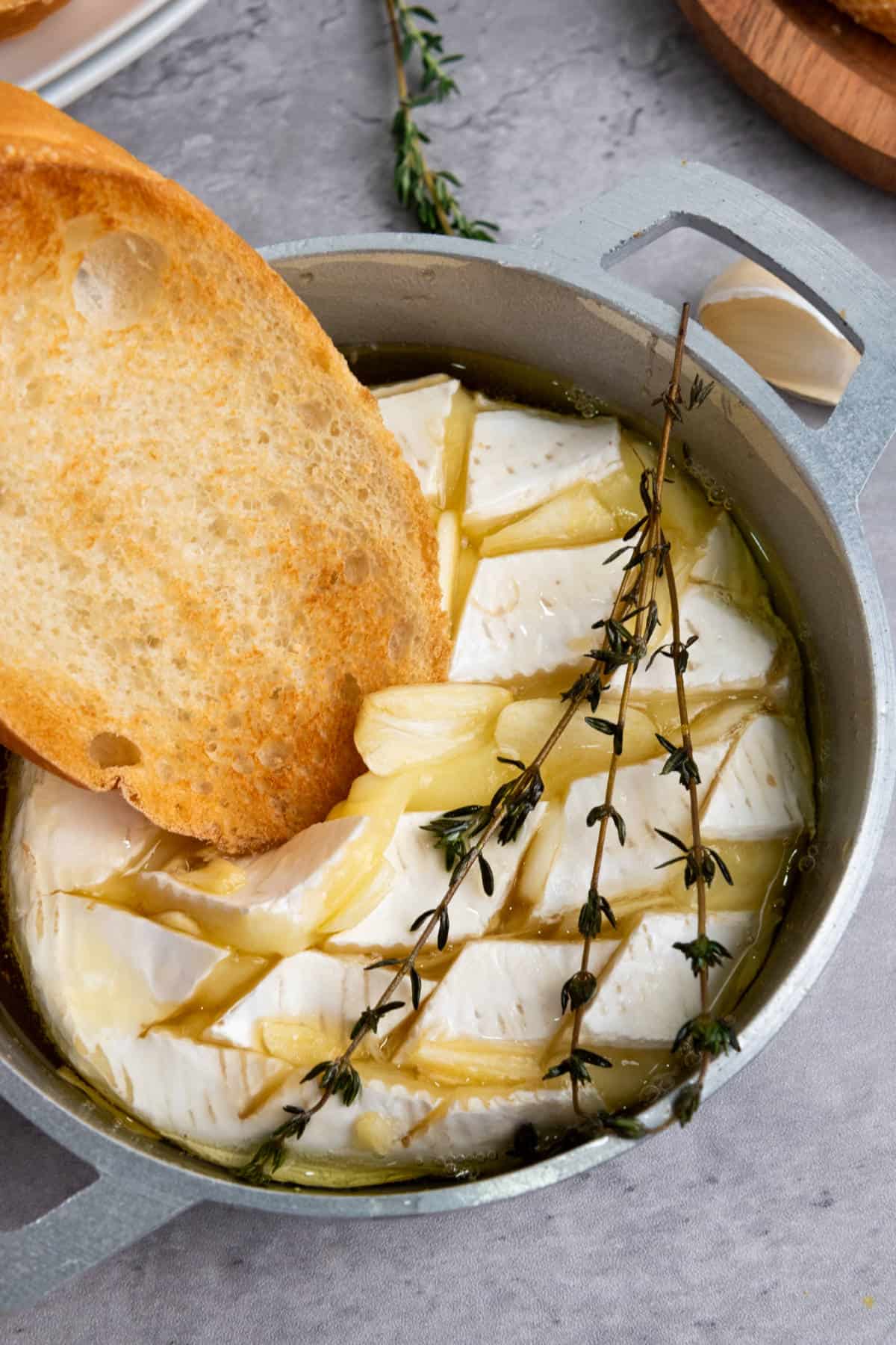 A round baked brie with honey, topped with olive oil, garlic slices, and thyme sprigs in a small pan. A toasted bread slice is dipped into the gooey melted cheese.