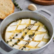A round baking dish with scored baked brie with honey, topped with fresh thyme and olive oil, surrounded by slices of toasted bread, garlic, and a wooden plate on a gray surface.