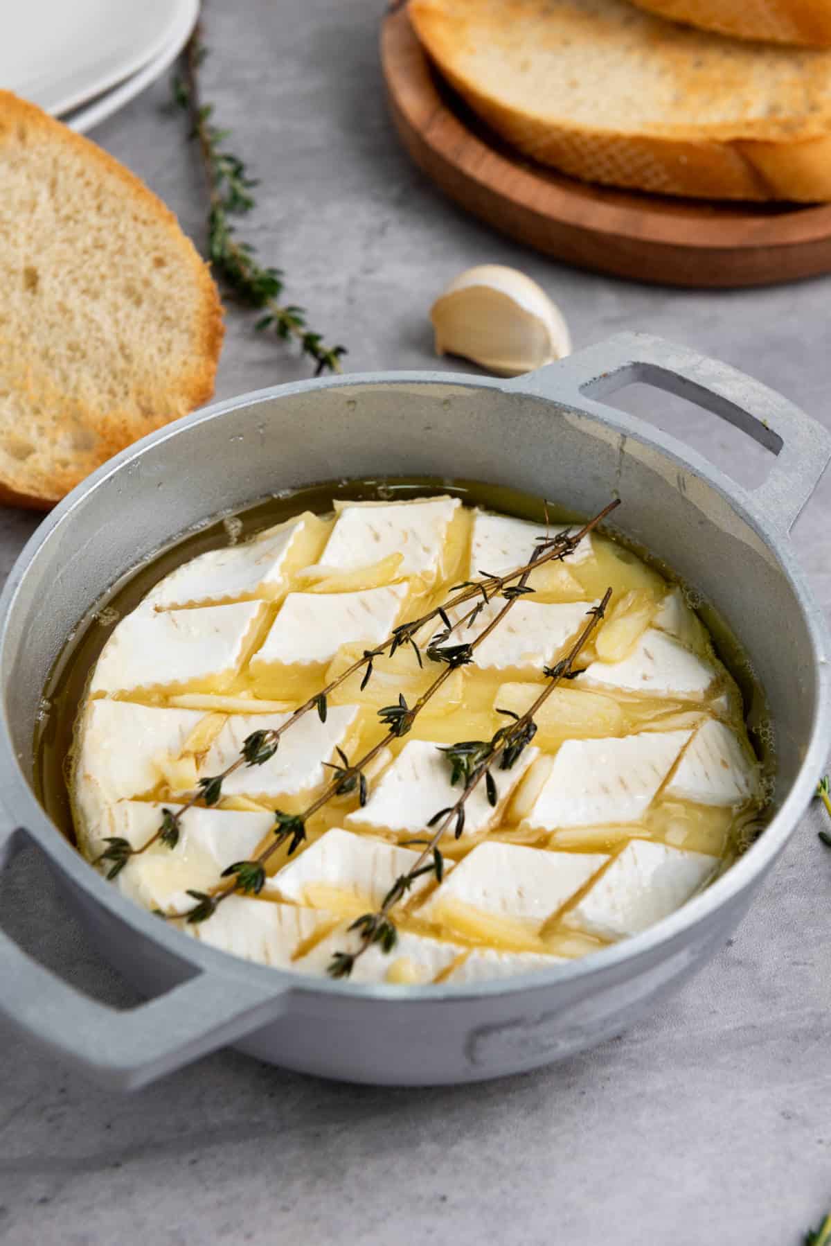 A round baking dish with scored baked brie with honey, topped with fresh thyme and olive oil, surrounded by slices of toasted bread, garlic, and a wooden plate on a gray surface.