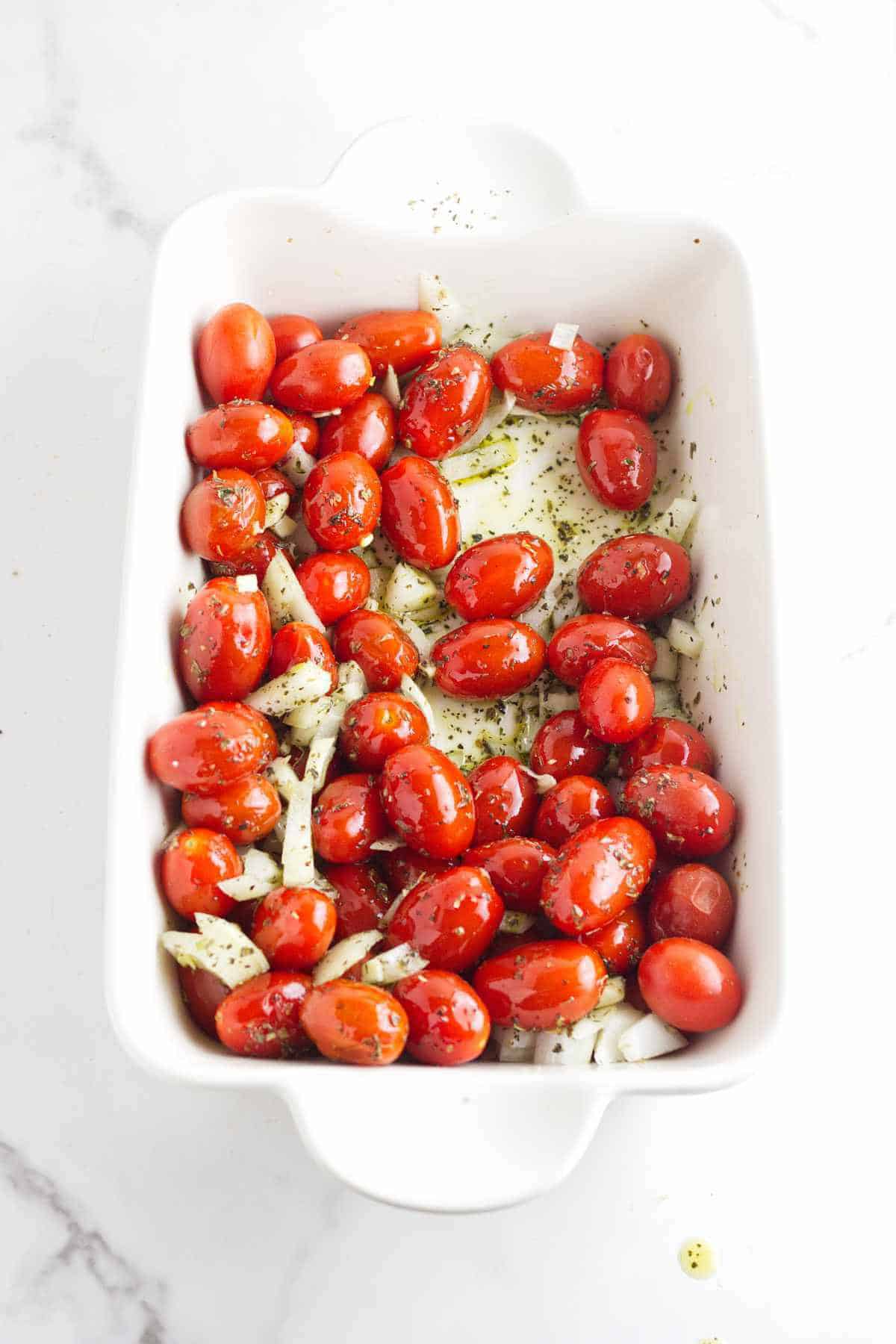A white baking dish filled with whole grape tomatoes, chopped onions, and sprinkled herbs-perfect for starting a flavorful feta cheese and tomato pasta-sits ready to be roasted on a white marble surface.