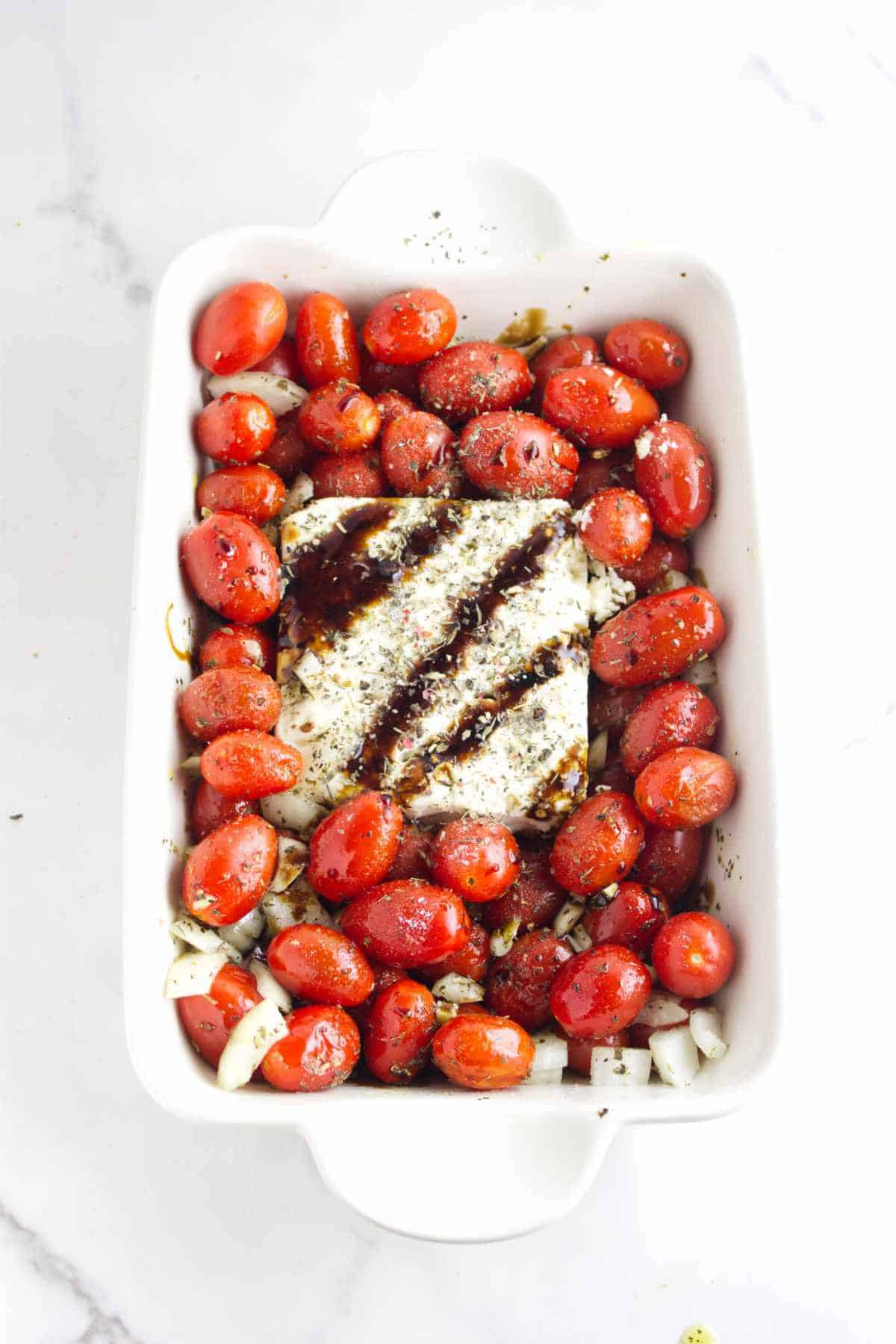A white baking dish filled with cherry tomatoes, diced onions, and a block of feta cheese, ready to become a delicious feta cheese and tomato pasta, topped with balsamic glaze and herbs on a white marble surface.