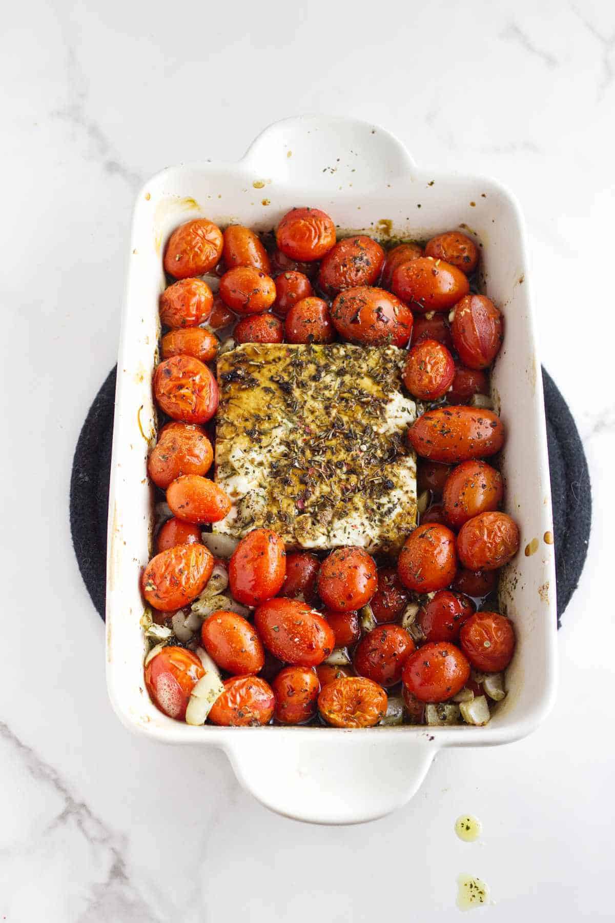 A white baking dish filled with roasted cherry tomatoes and a block of feta cheese in the center, sprinkled with herbs, sits on a white marble surface-perfect for making creamy feta cheese and tomato pasta.