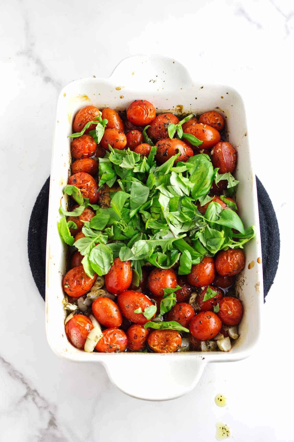 A white baking dish filled with roasted cherry tomatoes, chopped onions, and fresh basil leaves sits on a white marble surface-perfect for tossing into your favorite feta cheese and tomato pasta.