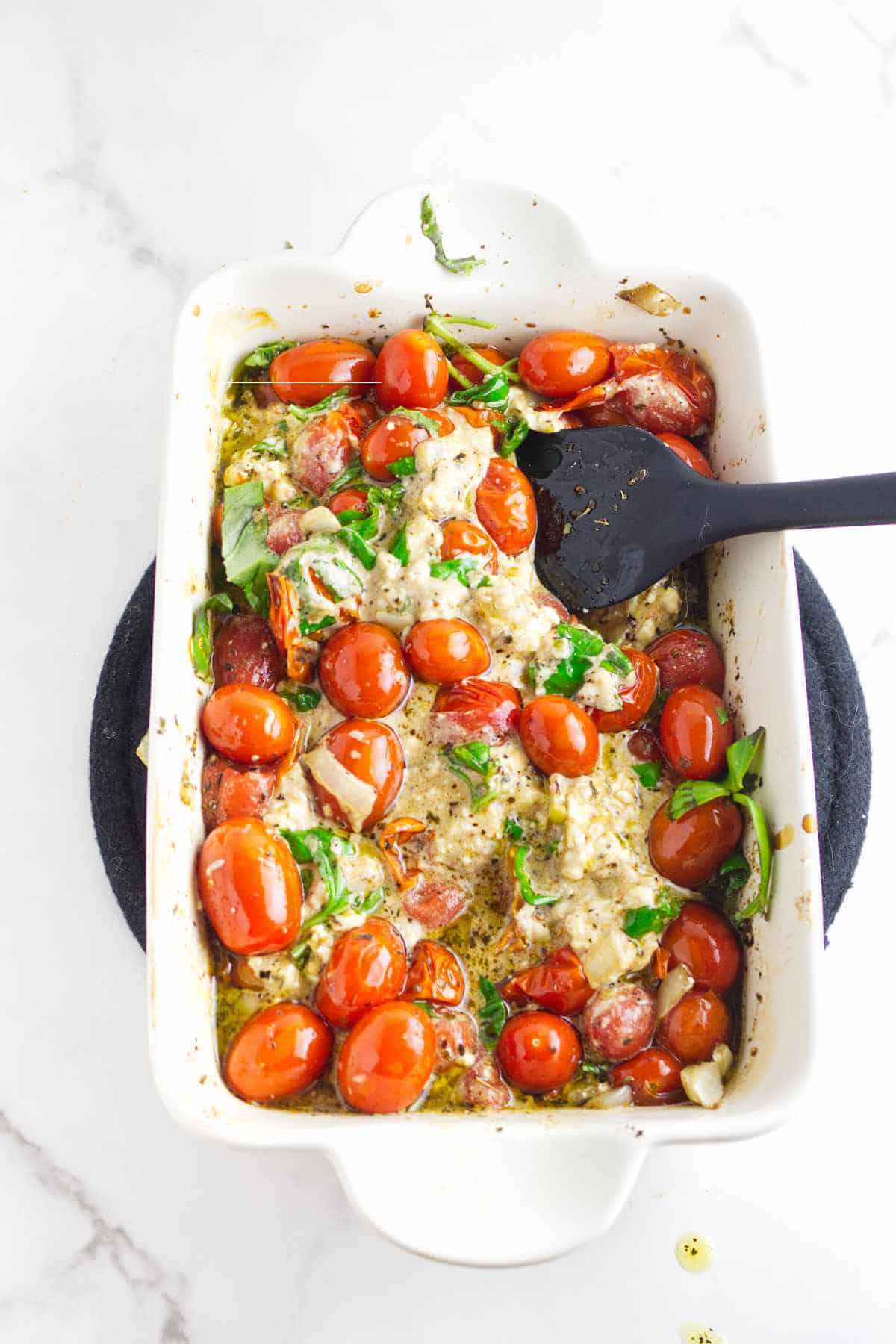 A white baking dish filled with creamy feta cheese and tomato pasta, whole cherry tomatoes, and fresh green herbs, with a black serving spoon resting inside. The dish sits on a dark round trivet on a white surface.