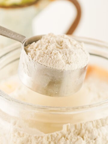 A metal measuring cup filled with flour hovers over an open glass jar of homemade bulk pancake mix, with more mix visible inside the jar. The background is softly blurred.