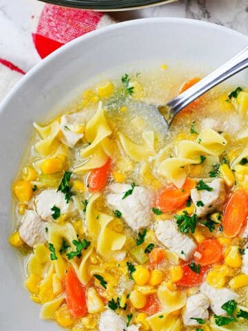 A bowl of chicken corn soup with carrots, egg noodles, and parsley, served with a spoon on a white plate beside a red and white checkered napkin. Corn adds sweetness to this comforting classic.