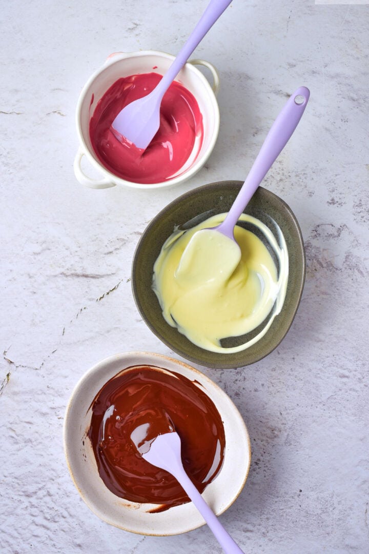 Three bowls of melted chocolate-pink, white, and dark-each with a purple spatula, are arranged vertically on a light marble surface, ready for making chocolate covered strawberries.