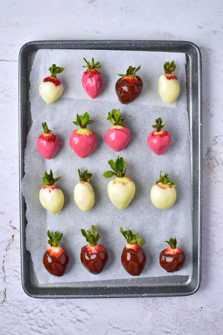 A baking tray lined with parchment paper holds chocolate covered strawberries dipped in white, pink, and dark chocolate, arranged in neat rows.