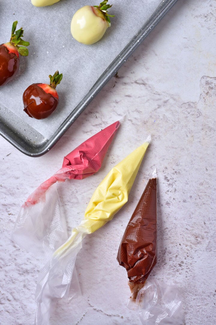 Piping bags filled with pink, yellow, and brown chocolate rest on a marble surface beside a tray of chocolate covered strawberries dipped in white and milk chocolate.