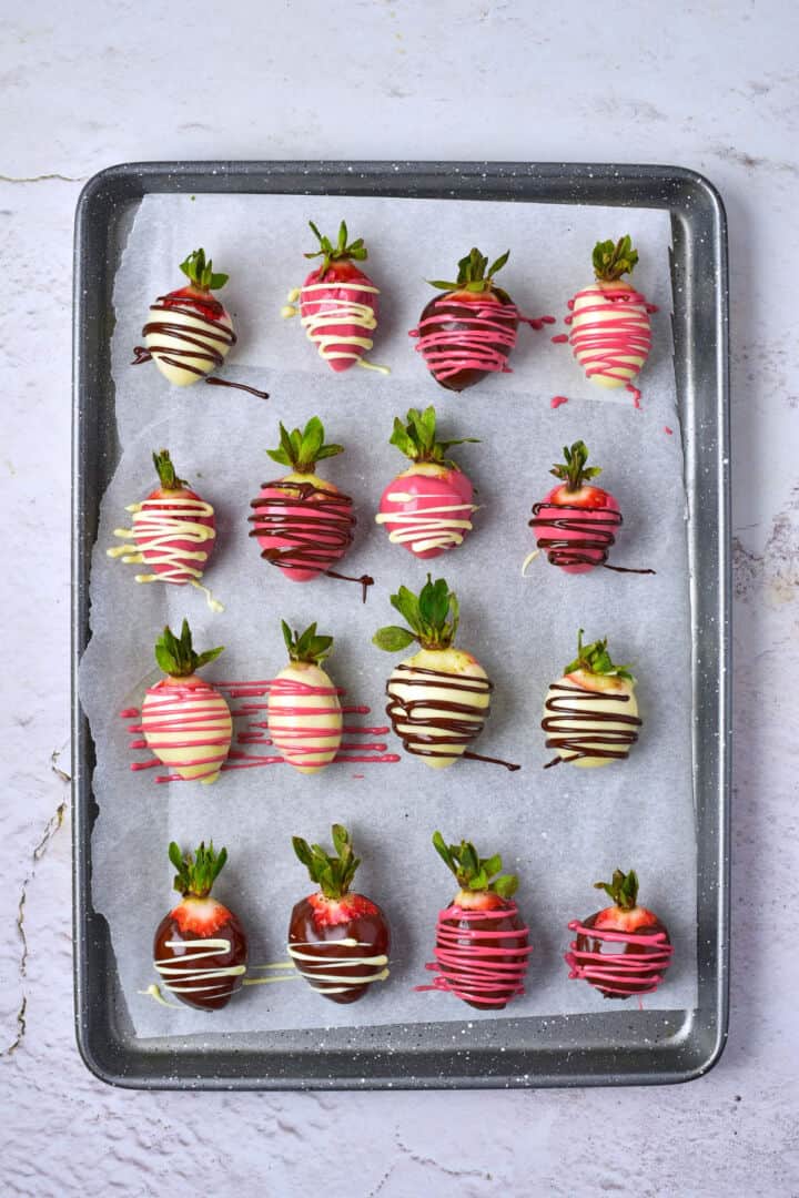 A tray of chocolate covered strawberries dipped in white, pink, and dark chocolate, each adorned with colorful chocolate drizzle and neatly arranged in rows on parchment paper.