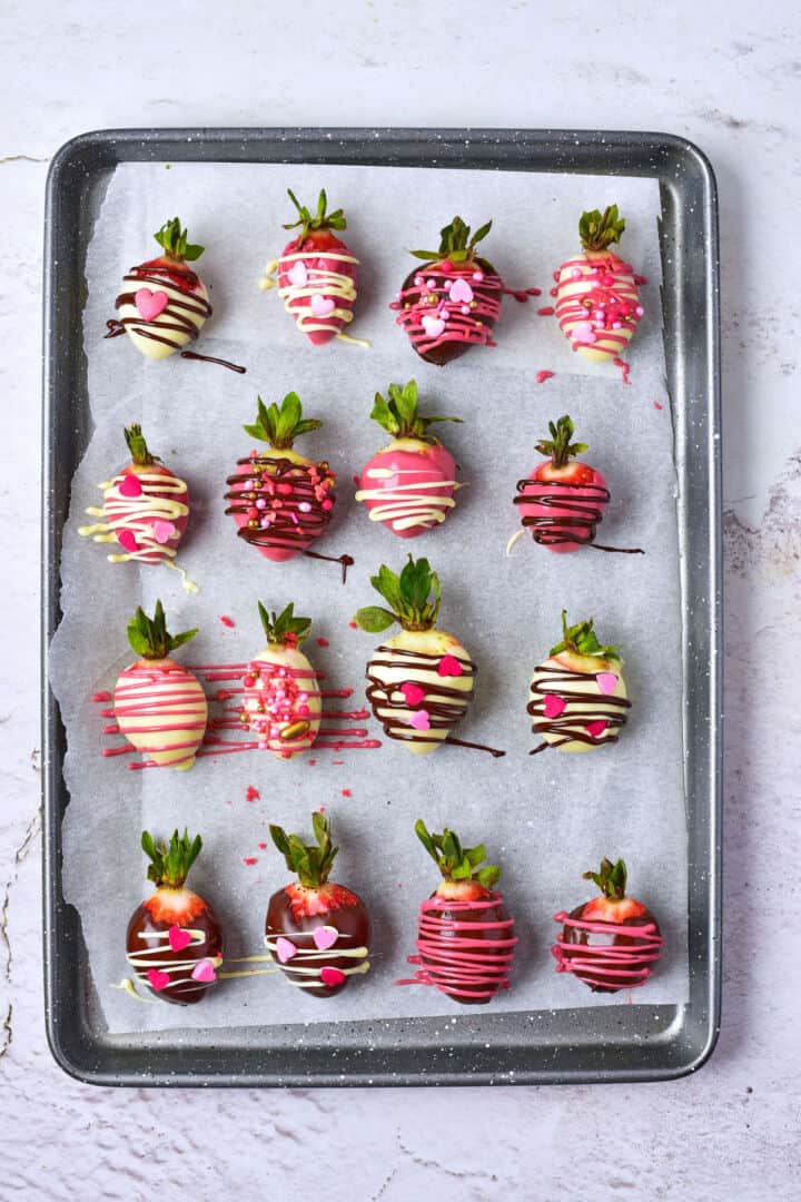A baking tray lined with parchment paper holds 15 chocolate covered strawberries, each dipped in chocolate and decorated with colorful drizzle, sprinkles, and heart-shaped toppings.