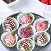 A round tray holds seven chocolate covered strawberries dipped in white and pink chocolate, decorated with drizzle, sprinkles, and heart-shaped toppings, each placed in a white paper cup. Bowls of chocolate and strawberries are nearby.