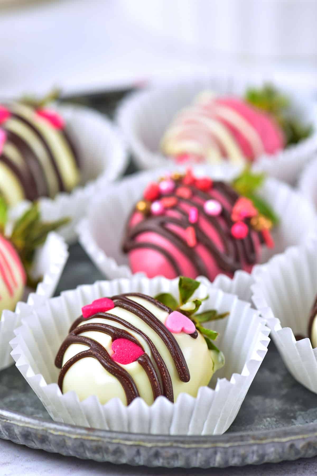 Chocolate covered strawberries decorated with white, pink, and dark chocolate drizzle and heart-shaped sprinkles, arranged in white paper cups on a metal tray.