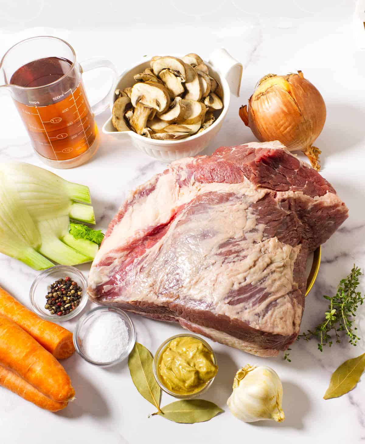 A large raw cider braised beef brisket surrounded by sliced mushrooms, an onion, fennel bulb, carrots, broth, garlic, mustard, bay leaves, salt, peppercorns, and thyme sprigs on a white surface.