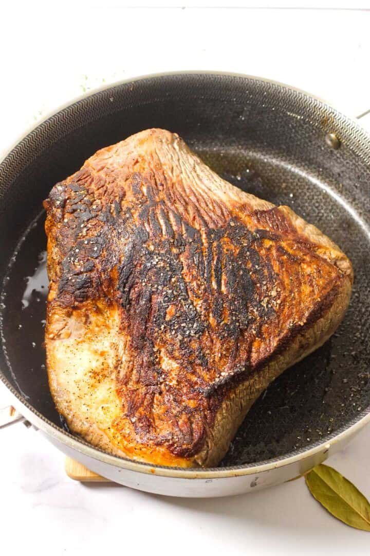 A large, browned piece of cider braised beef brisket is searing in a black frying pan. The meat has a well-seared crust and is sprinkled with salt and pepper. A bay leaf sits beside the pan on a white surface.