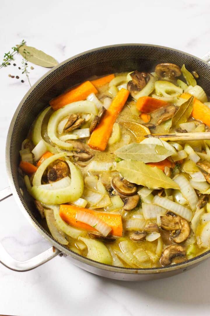 A pot filled with sliced onions, carrots, mushrooms, and bay leaves simmering in broth alongside tender cider braised beef brisket, with fresh herbs visible on the side.