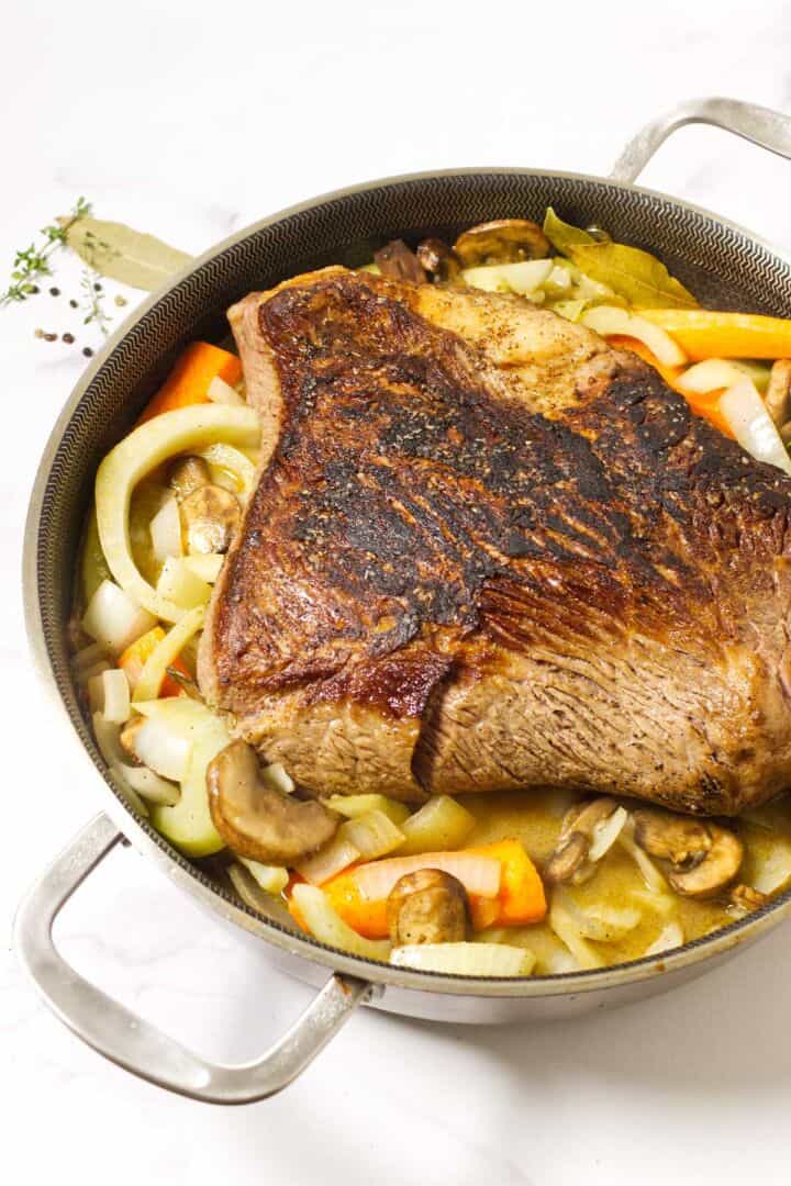 A browned cider braised beef brisket sits atop sliced onions, carrots, celery, and mushrooms in a large metal pan, ready for roasting. Fresh herbs and peppercorns are visible nearby on a white surface.
