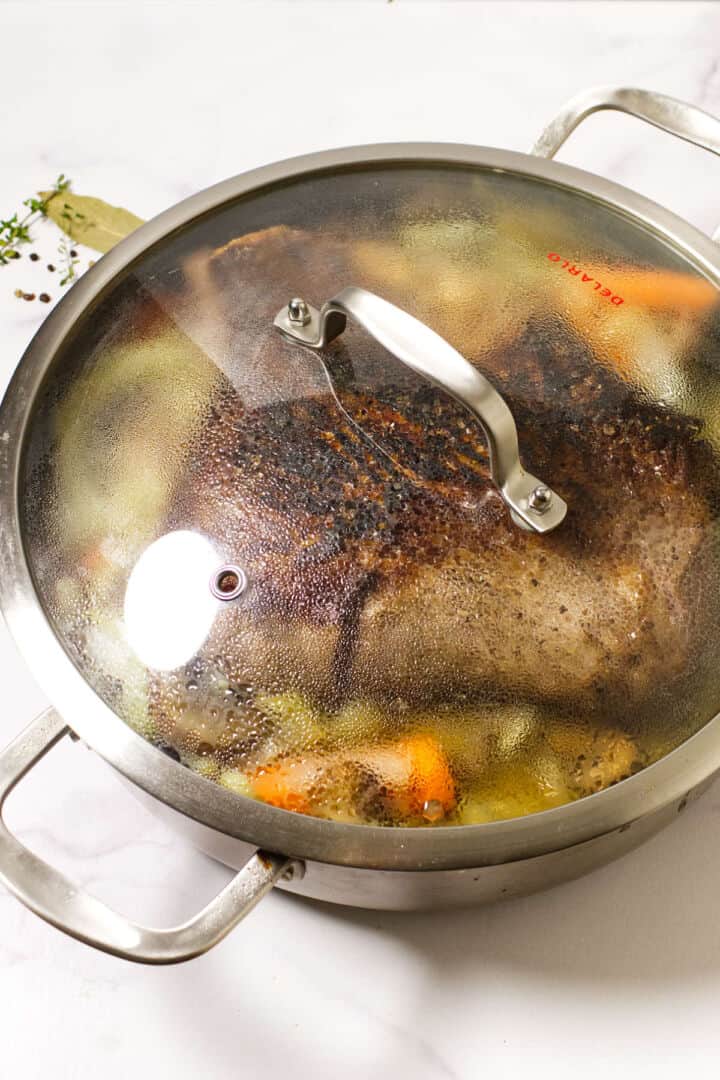 A large metal pot with a glass lid shows a cider braised beef brisket, cooked to perfection and surrounded by browned carrots and vegetables. Steam and condensation cover the lid, while herbs rest outside the pot on a white surface.
