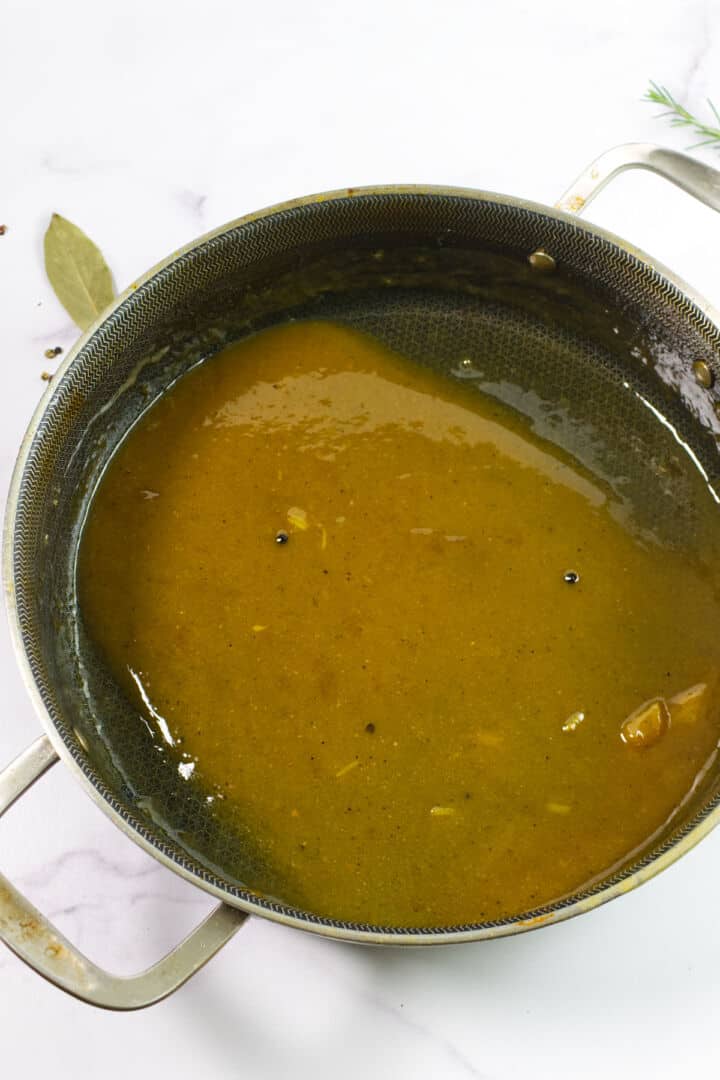 A metal saucepan filled with smooth brown gravy sits on a white countertop. A bay leaf and some herbs are visible nearby, suggesting the gravy was freshly made and seasoned for cider braised beef brisket.