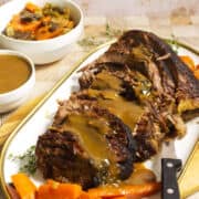 A platter of cider braised beef brisket topped with brown gravy, served with cooked carrots. A carving knife rests beside the roast. In the background are bowls of extra gravy and mixed vegetables.