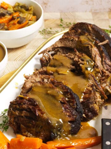 A platter of cider braised beef brisket topped with brown gravy, served with cooked carrots. A carving knife rests beside the roast. In the background are bowls of extra gravy and mixed vegetables.