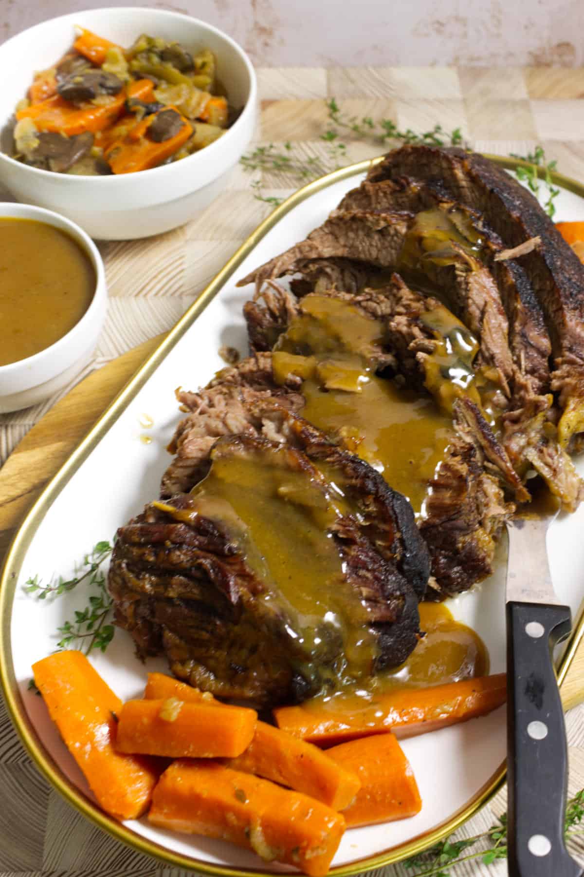 A platter of sliced cider braised beef brisket topped with brown gravy, served with cooked carrots. A knife rests on the plate. In the background are bowls of extra gravy and mixed cooked vegetables.
