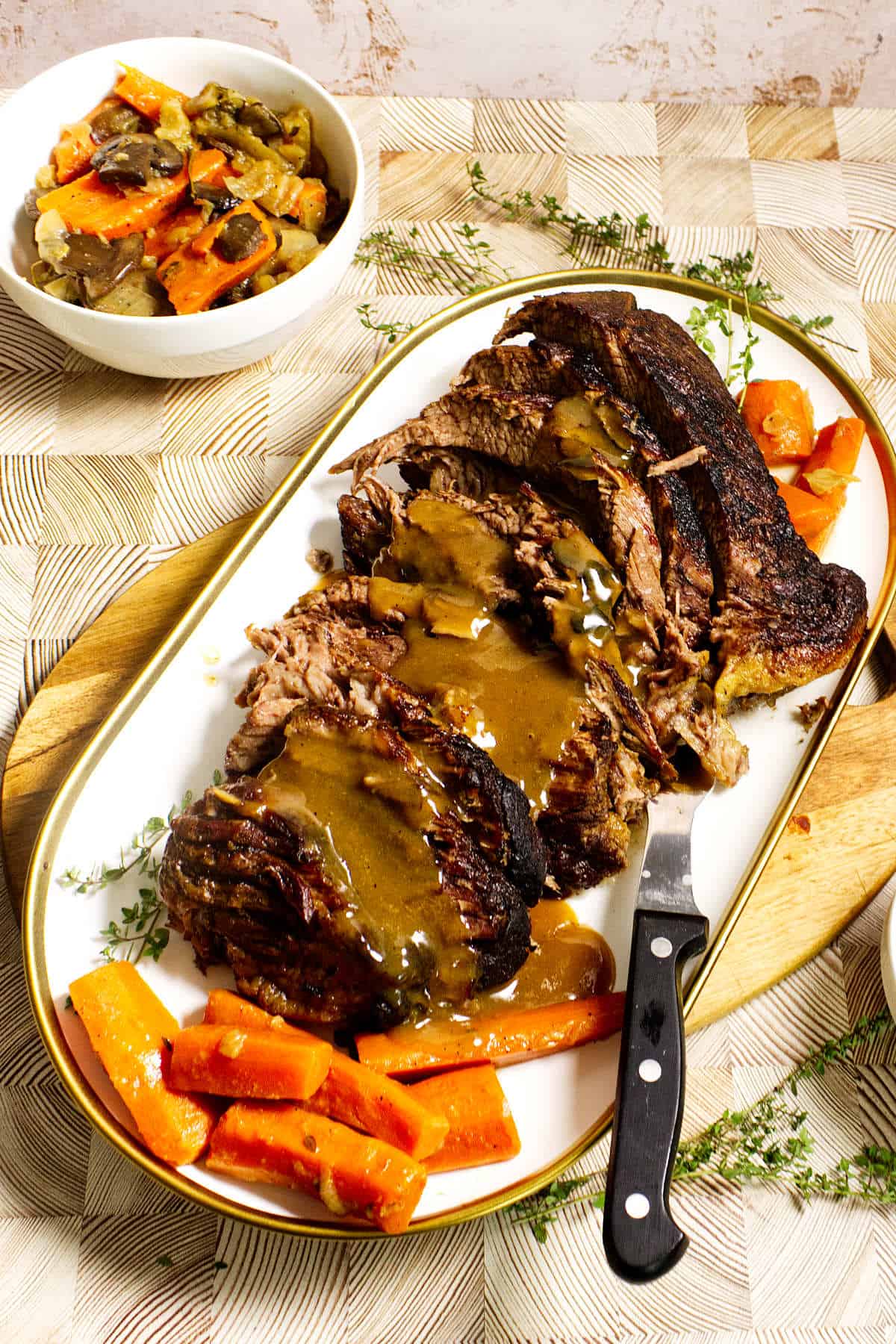 Sliced cider braised beef brisket topped with brown gravy on a white platter, garnished with cooked carrots and fresh herbs. A serving knife rests on the platter, with a bowl of roasted vegetables visible in the background.