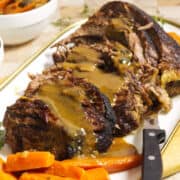 A sliced cider braised beef brisket topped with brown gravy, served on a white platter with cooked carrots and a knife. A bowl of vegetables is visible in the background.