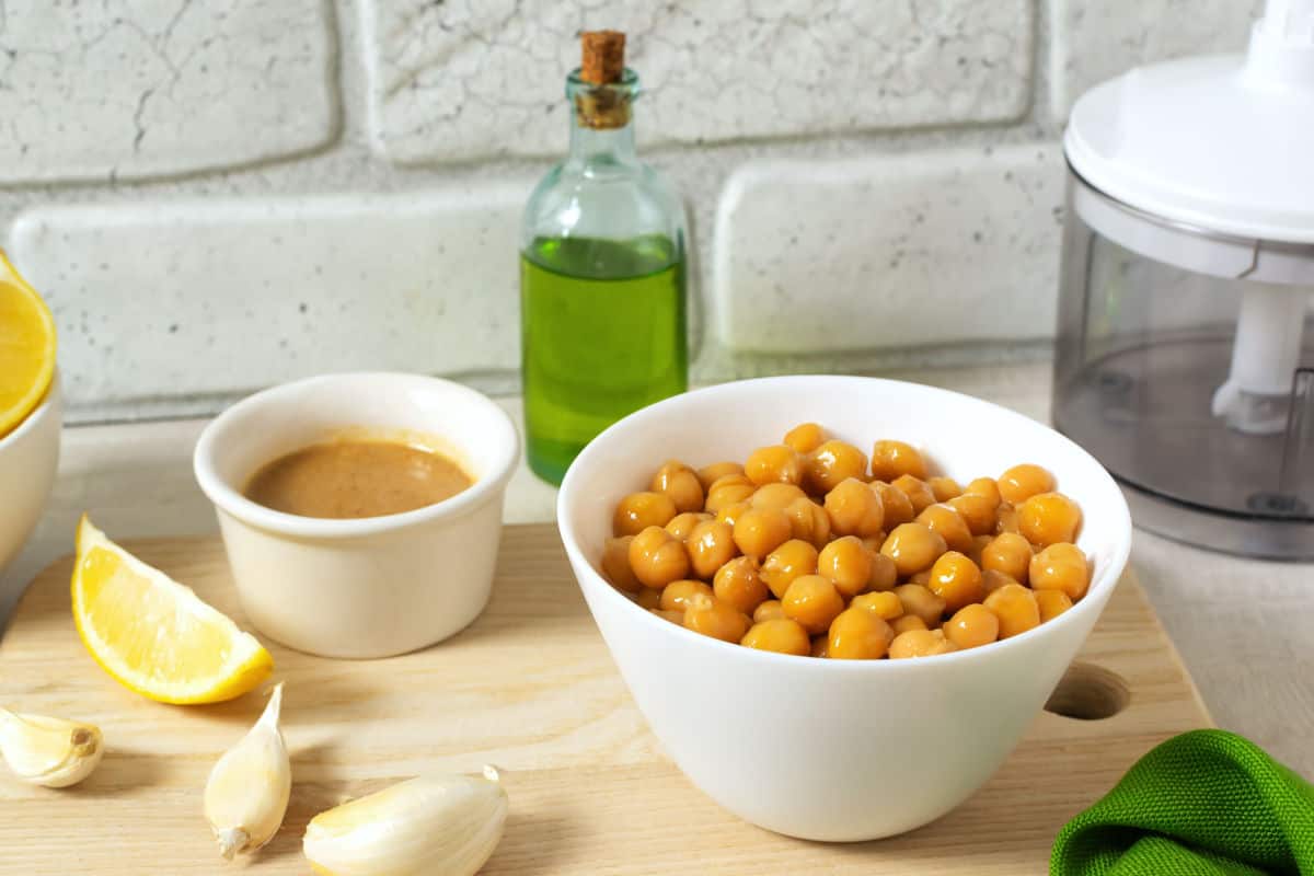 A white bowl of chickpeas, a small bowl of tahini, garlic cloves, a lemon wedge, and a bottle of olive oil are arranged on a wooden surface—perfect ingredients for making classic hummus dip with a food processor in the background.
