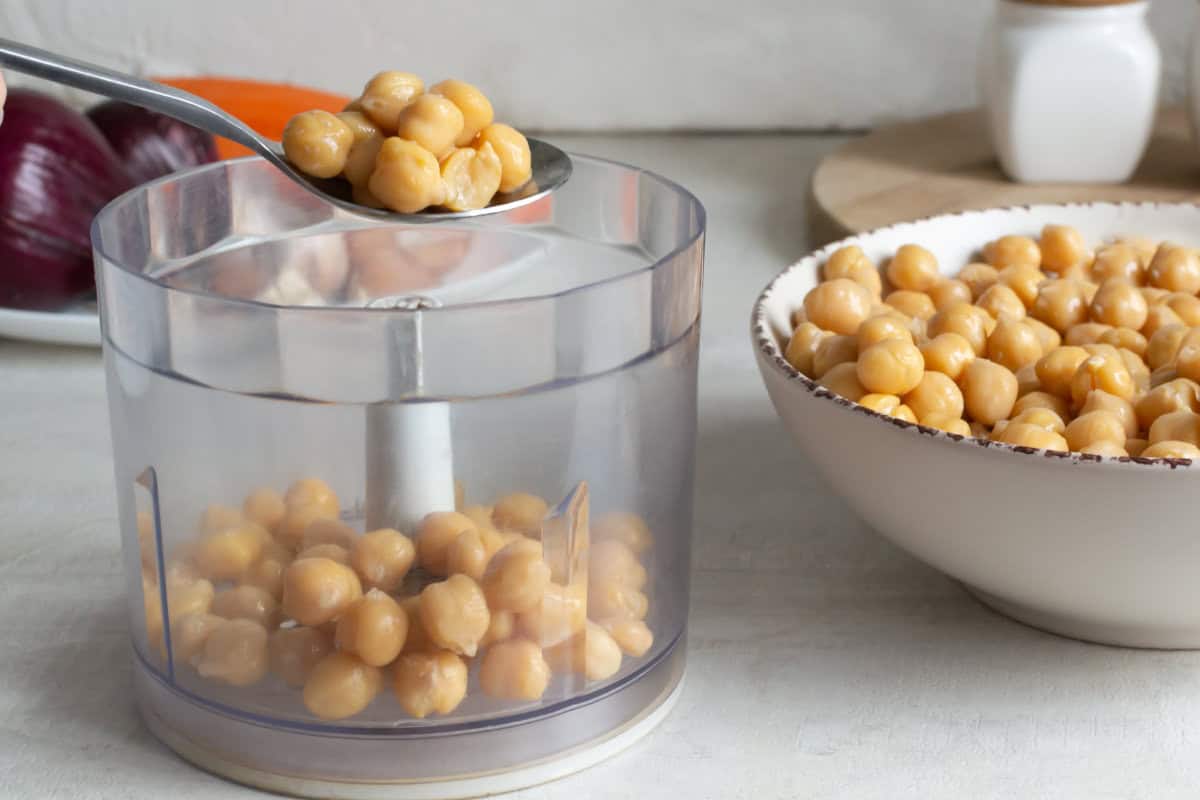 A spoon holds chickpeas over a food processor bowl partially filled with chickpeas, ready to be blended into homemade hummus dip. A large white bowl of chickpeas sits nearby, with red onions and kitchen items blurred in the background.