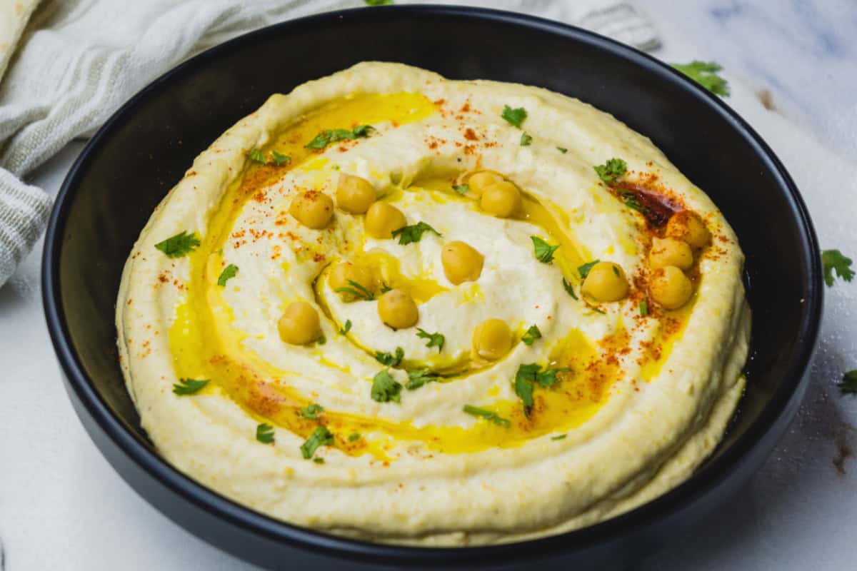 A black bowl filled with creamy hummus dip, garnished with whole chickpeas, a drizzle of olive oil, chopped parsley, and a sprinkle of paprika.