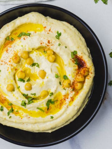 A black bowl filled with creamy hummus dip, topped with whole chickpeas, a drizzle of olive oil, chopped parsley, and a sprinkle of paprika. The bowl sits on a white surface with parsley scattered around.